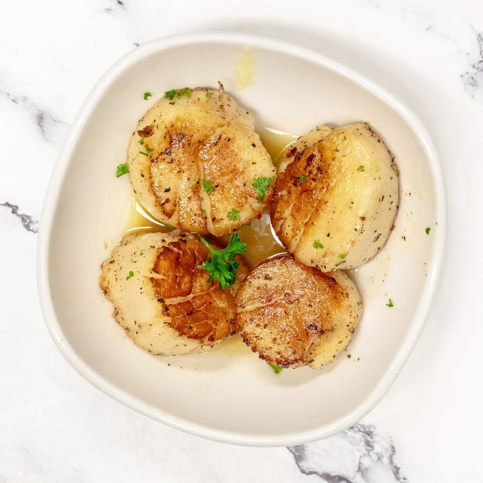 Four seared scallops with a golden-brown crust, garnished with fresh parsley in a white bowl.