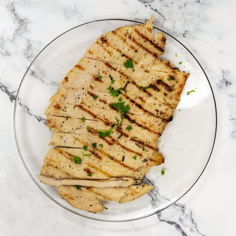 Grilled chicken breast, sliced and garnished with parsley, on a clear plate.