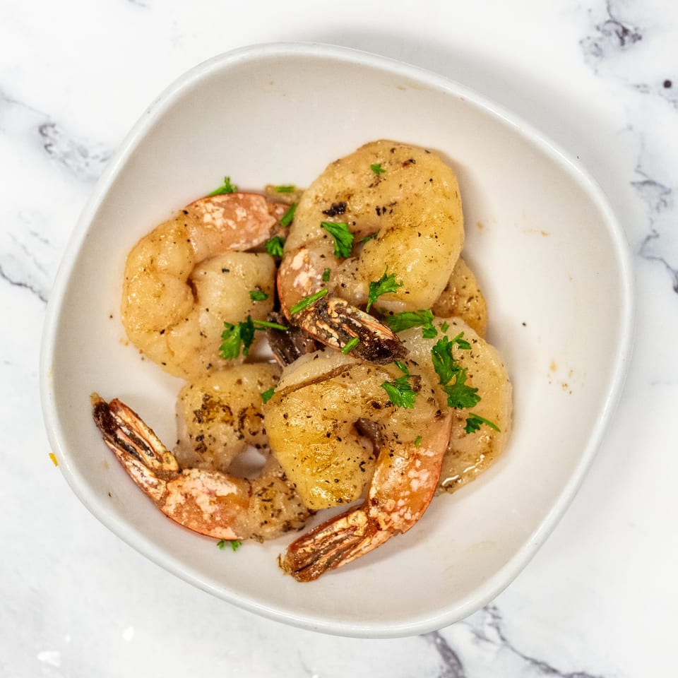 A white bowl filled with seasoned shrimp garnished with fresh parsley on a marble surface.