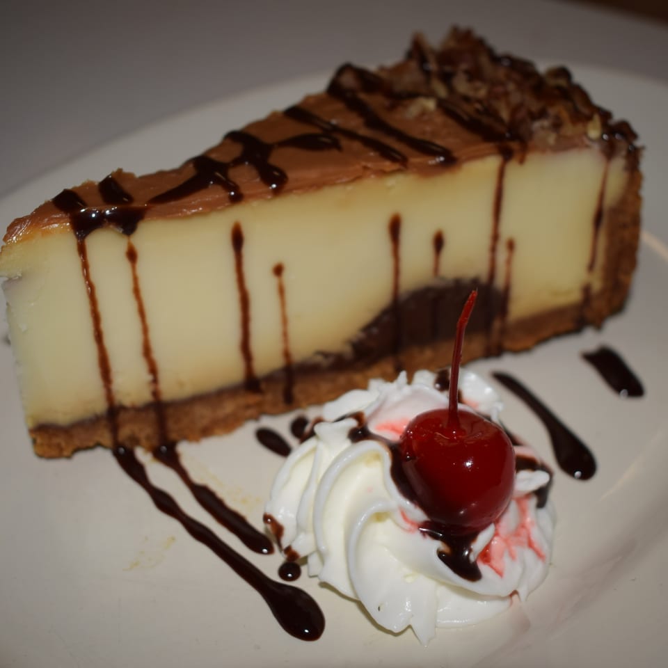 Slice of cheesecake with chocolate drizzle, whipped cream, and a cherry on a white plate.