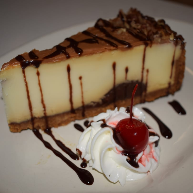 Slice of cheesecake with chocolate drizzle, whipped cream, and a cherry on a white plate.
