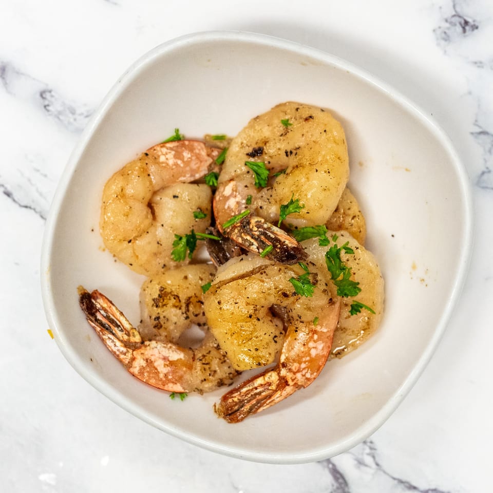 A white bowl filled with seasoned shrimp garnished with fresh parsley on a marble surface.