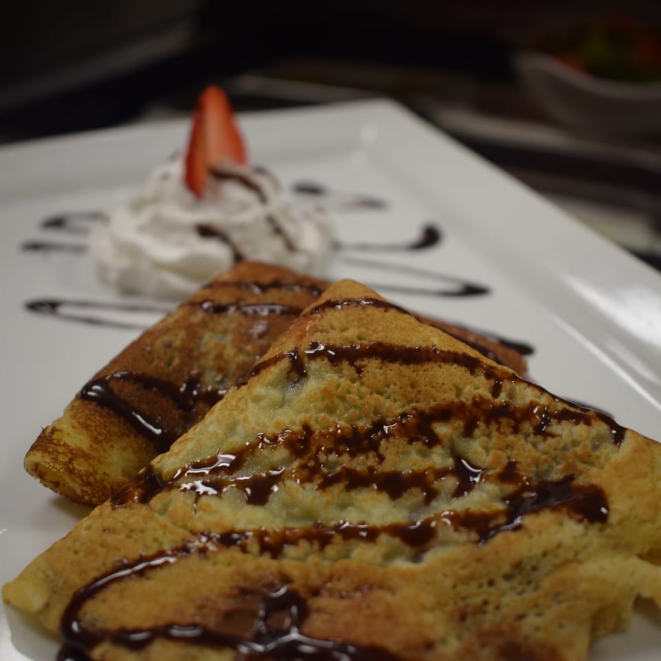 Two golden crepes drizzled with chocolate sauce, topped with whipped cream and a strawberry slice.